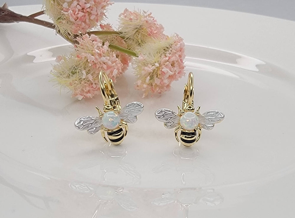 Gold bee-shaped earrings with opal stones on a white surface with pink flowers.
