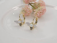 Gold bee-shaped earrings on a white plate with pink flowers in the background