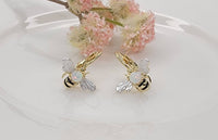 Gold bee-shaped earrings with opal stones on a white surface with pink flowers.