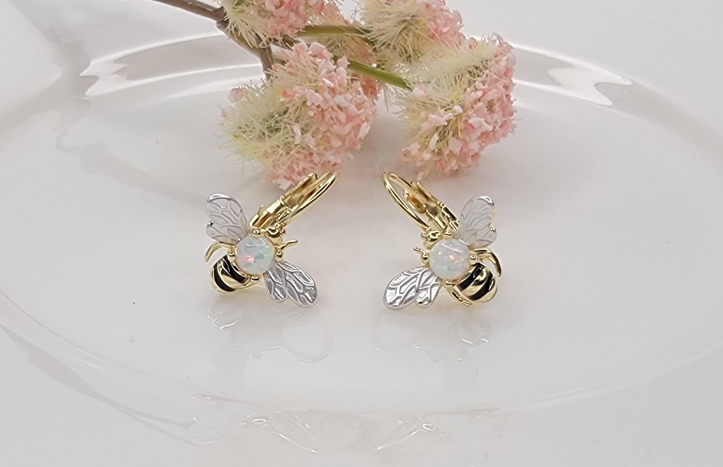 Gold bee-shaped earrings with opal stones on a white surface with pink flowers.