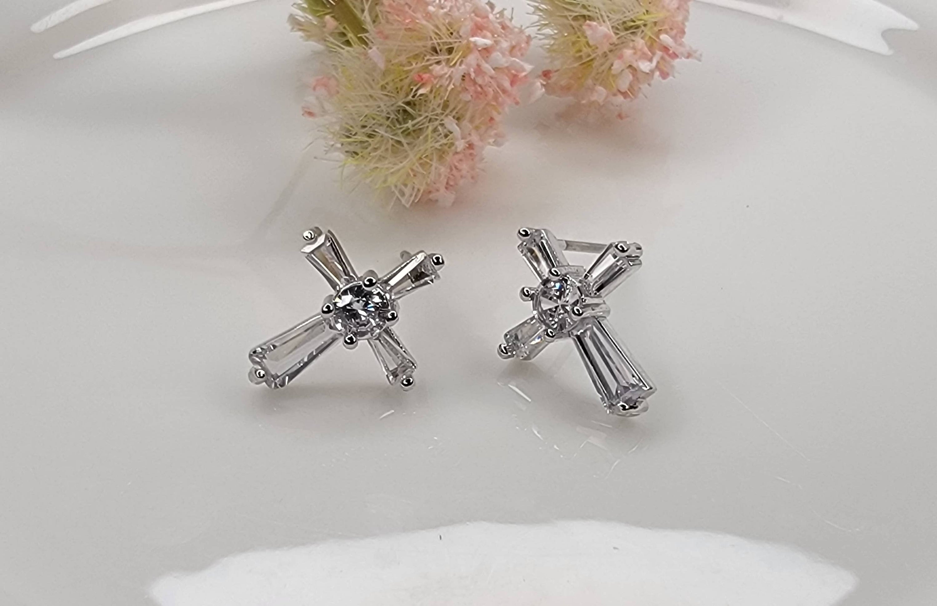 Silver cross-shaped earrings on a white surface with pink flowers in the background