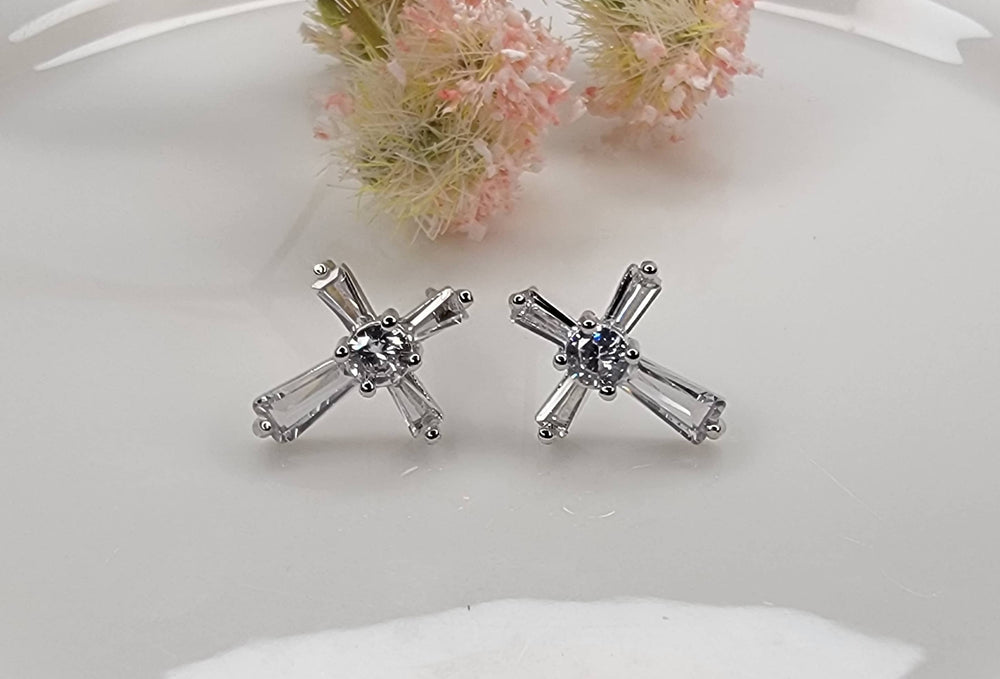 Silver cross-shaped earrings on a white surface with pink flowers in the background